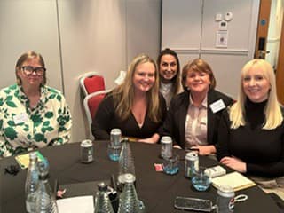 Five people smiling at a table with drinks and notepads in a conference setting.