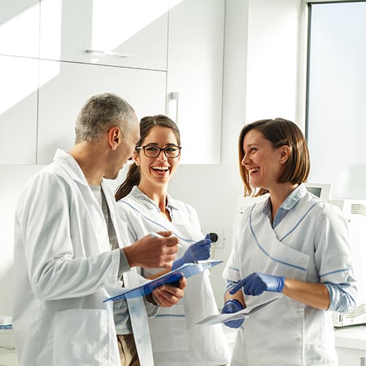 Three dentists in white coats laughing, holding clipboards and papers.