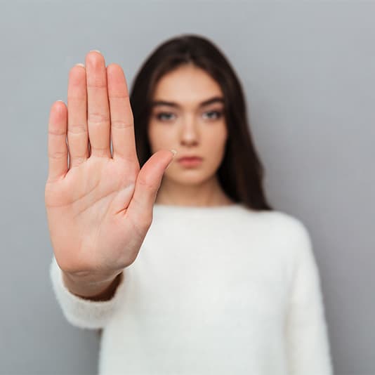 Woman in a white sweater holding her hand up towards the camera, palm open, in a gesture suggesting "stop" or refusal, against a grey background.