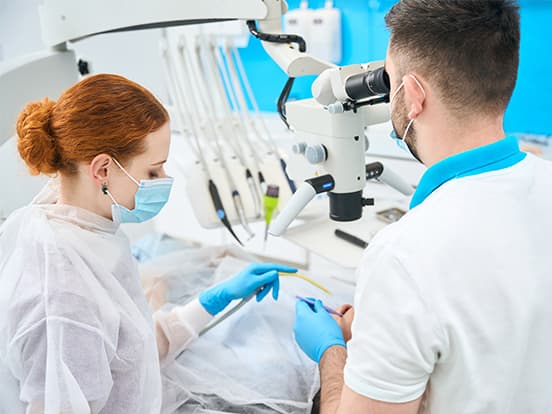 Two dental professionals, wearing masks and gloves, work on a patient using a dental microscope in a brightly lit clinic.