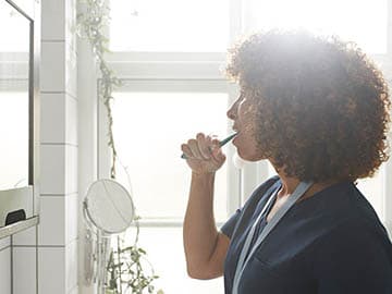 Person with curly hair brushing teeth in a bright bathroom, standing by a window with sunlight streaming in.