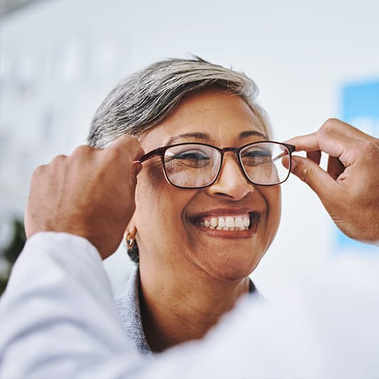 A smiling woman tries on glasses with the help of an optician in a bright room.