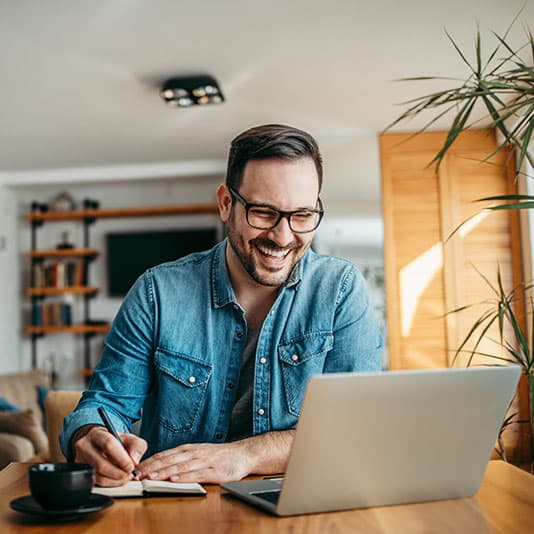 Man in glasses, wearing a denim shirt, smiling while writing in a notebook and using a laptop at a wooden table in a bright, modern room.