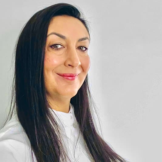 Smiling person with long dark hair, wearing a white top, against a plain light gray background.