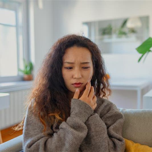 A woman with curly hair and a painful expression sits on a couch in a bright room, wearing a gray sweater.