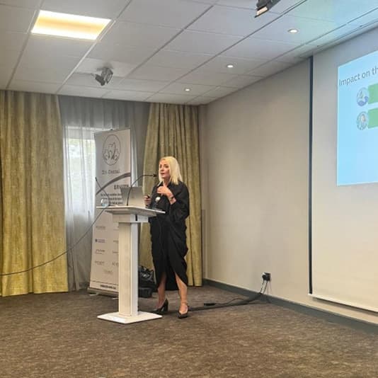 A woman stands at a podium giving a presentation in a conference room with a projector screen displaying a slide.