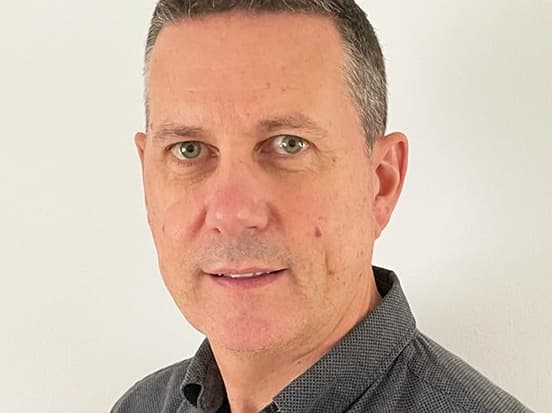 A man with short dark hair and a light complexion, wearing a dark collared shirt, looks at the camera against a plain white background.