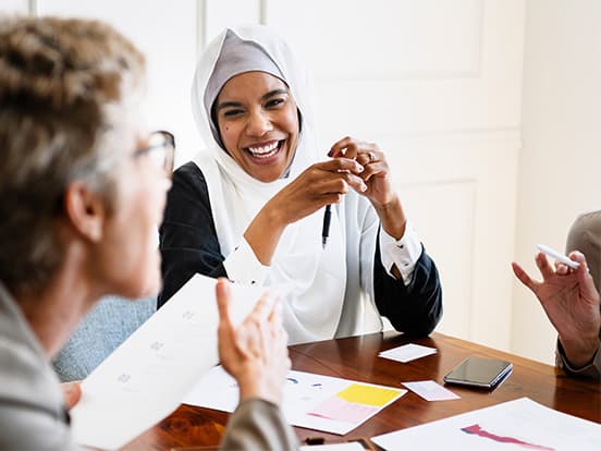 Three people at a meeting table, smiling and discussing papers, with one woman wearing a hijab and holding a pen.