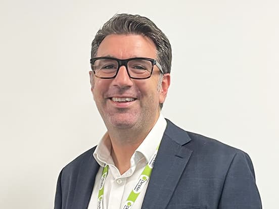 Smiling man in a suit with glasses, wearing a lanyard, standing against a plain white background.