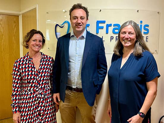 Three people standing and smiling in an office with a dental practice sign in the background.